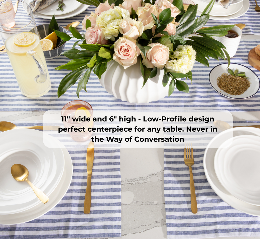 A styled table setting featuring a white ribbed vase filled with pink and cream roses and green leaves, placed on a blue and white striped tablecloth. White plates, gold cutlery, a glass pitcher with lemon, and small bowls are arranged around it.