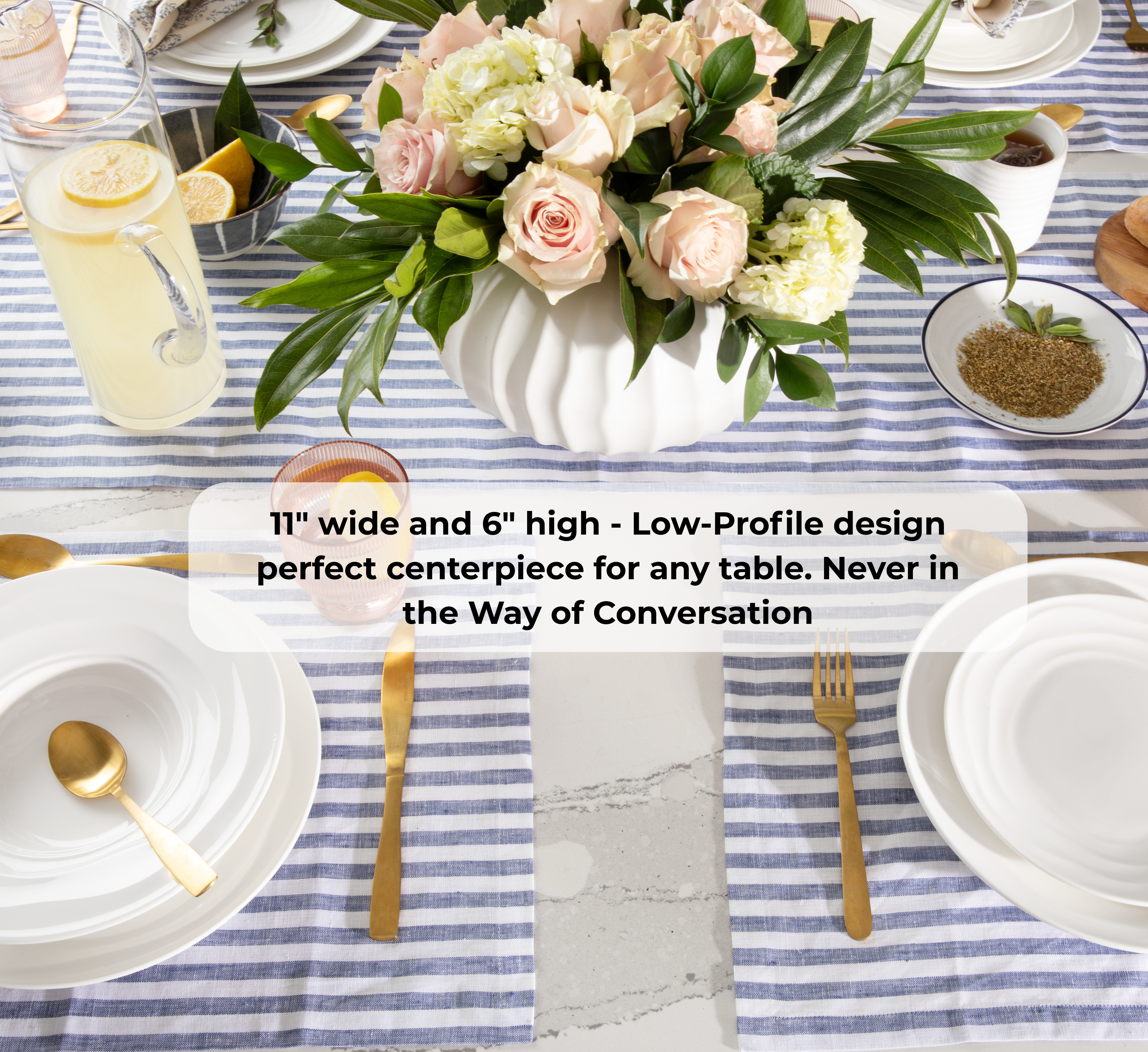 A styled table setting featuring a white ribbed vase filled with pink and cream roses and green leaves, placed on a blue and white striped tablecloth. White plates, gold cutlery, a glass pitcher with lemon, and small bowls are arranged around it.