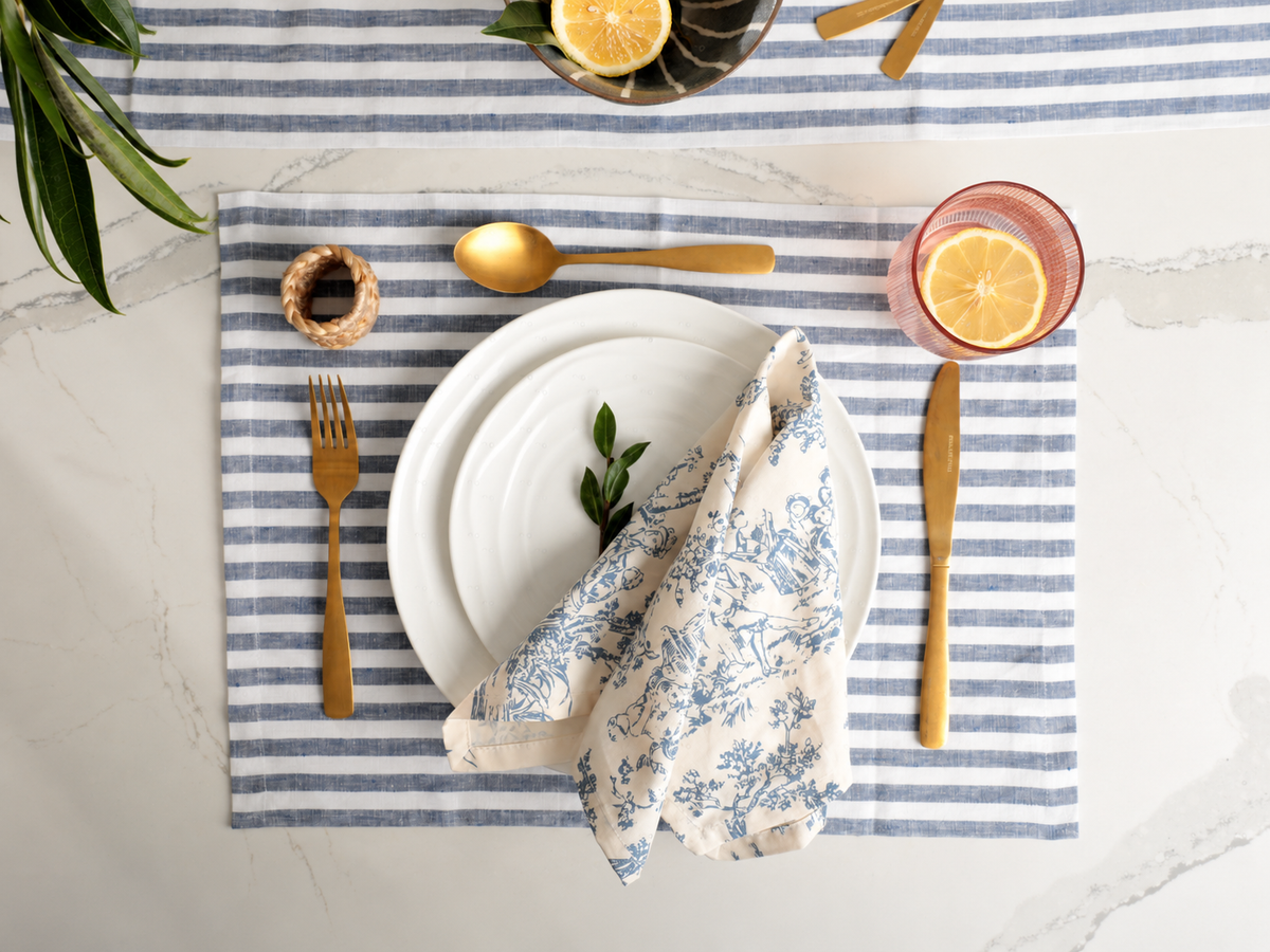 Overhead flat-lay of a table setting with Natural Waves plates, gold flatware, striped linen placemat, and citrus garnish